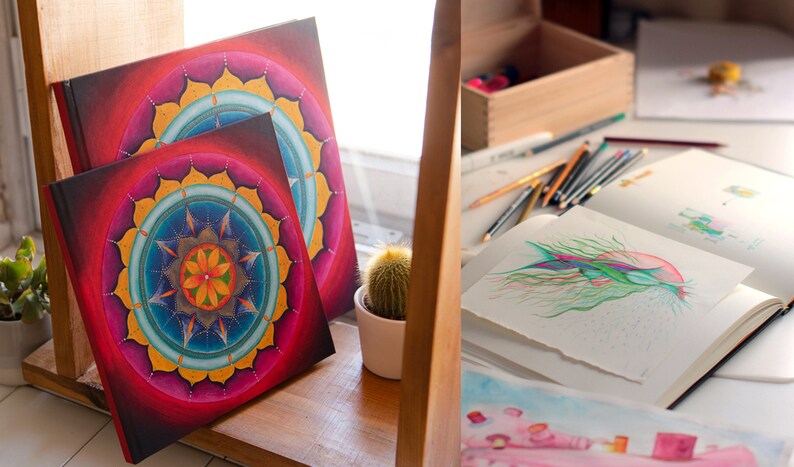 May include: Two square journals with colorful mandala designs on the covers. The journals are stacked on a wooden shelf with a small cactus plant in a white pot.
