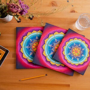 May include: Three colorful mandala notebooks with pink, yellow, blue, and green designs. The notebooks are stacked on a wooden surface.