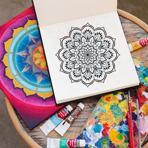 May include: An open sketchbook with a black and white mandala design on the right page. A painted mandala with bright colors is on the left page. A wooden table with paint tubes and a paint palette with colorful paint is in the foreground.