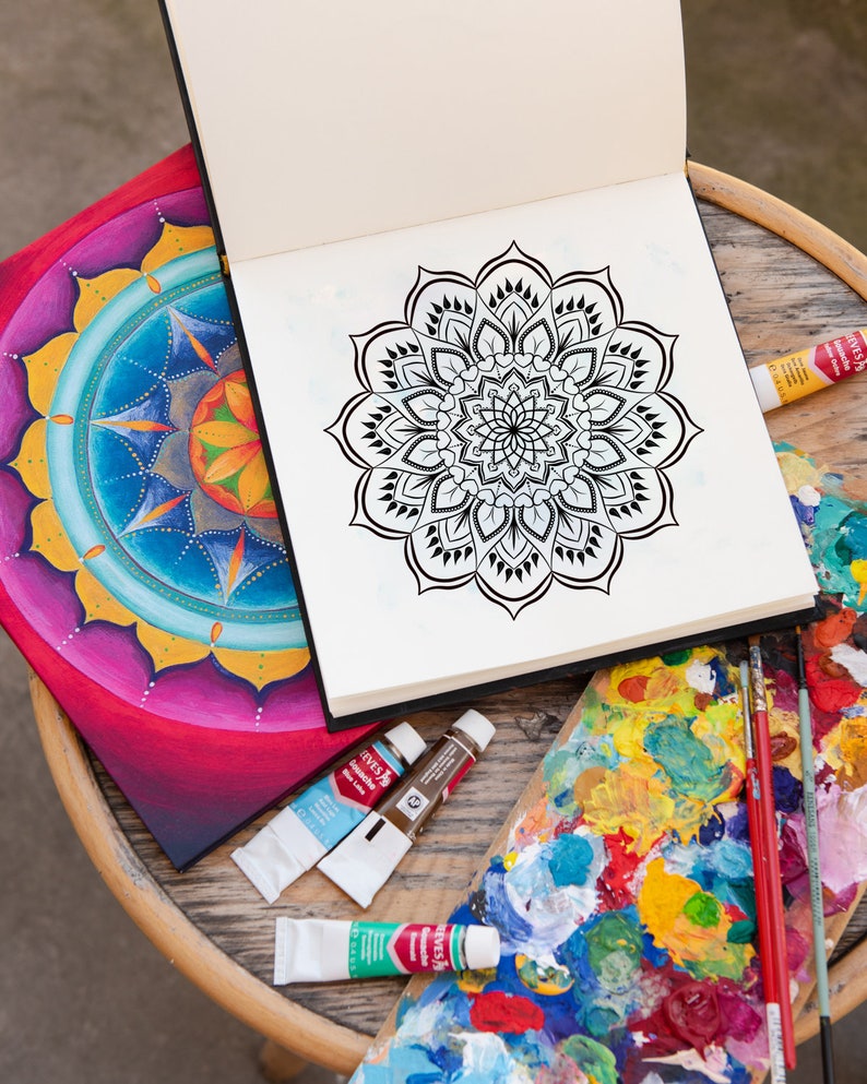 May include: An open sketchbook with a black and white mandala design on the right page. A painted mandala with bright colors is on the left page. A wooden table with paint tubes and a paint palette with colorful paint is in the foreground.