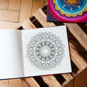 May include: An open coloring book with a black and white mandala design on the right page. The book is sitting on a wooden pallet. A painted mandala is visible in the background.