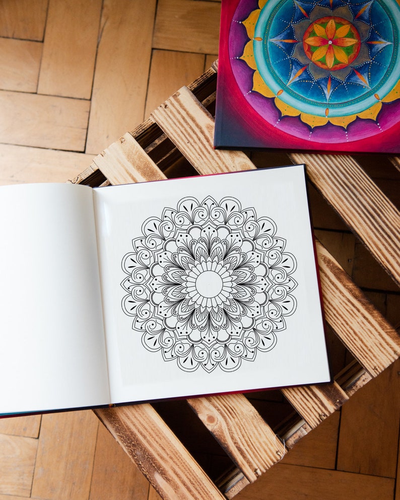 May include: An open coloring book with a black and white mandala design on the right page. The book is sitting on a wooden pallet. A painted mandala is visible in the background.