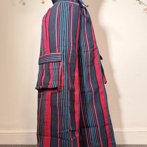 May include: Wide-leg cargo trousers featuring a vertical striped pattern in dark blue, bright red, and light blue. The trousers have an elastic waistband with a drawstring tie and large rectangular cargo pockets on the sides of each leg.