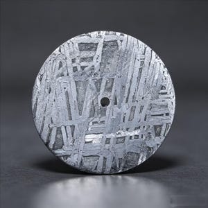 Puede incluir: Un disco metálico circular de meteorito Muonionalusta con un patrón de Widmanstätten distintivo de líneas cristalinas entrecruzadas y un pequeño orificio circular centrado. La superficie está pulida, mostrando tonos plateados y grises.