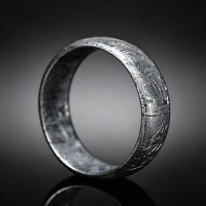 Puede incluir: Un anillo metálico con acabado gris oscuro y superficie texturizada que presenta líneas intrincadas y entrecruzadas, creando un patrón orgánico complejo en toda la banda. El anillo tiene un perfil liso y redondeado con un brillo pulido y reflectante.