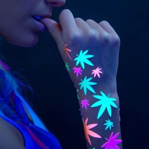 Könnte beinhalten: Eine Hand mit leuchtenden Neon-Blatt-Tattoos in Grün-, Orange- und Lilatönen. Die Tattoos sind auf dem Unterarm vor einem dunklen Hintergrund angeordnet. Das Bild wird mit Schwarzlicht beleuchtet.