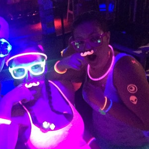 May include: Two people wearing neon glow-in-the-dark accessories, including glasses, necklaces, and bracelets. One person has a mustache and the other has a mustache and a pair of hands on their chest.