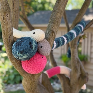 May include: A collection of crocheted plush snakes in various colours including cream, dark blue, light brown, and bright pink. The snakes feature soft, textured yarn bodies with small black bead eyes, coiled and draped across the branches of a tree.