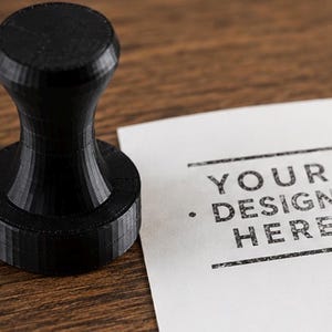 May include: A black circular hand stamp with a vertical handle. Next to the stamp is a white piece of paper featuring a black ink impression that reads: YOUR DESIGN HERE, framed by two horizontal lines and two small dots on either side.