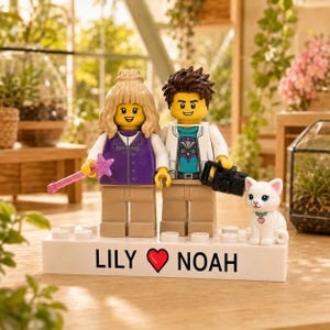 Puede incluir: Dos minifiguras LEGO, una con cabello rubio y chaleco morado sosteniendo una varita de estrella rosa, la otra con cabello castaño y chaqueta blanca sosteniendo una cámara negra, sobre una base blanca con el texto LILY (corazón) NOAH y un gato blanco.