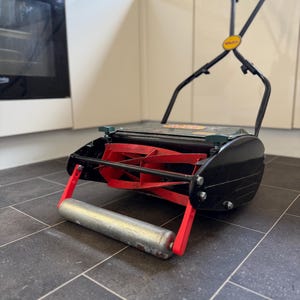 Op de afbeelding: Een handmatige grasmaaier met een zwart metalen frame, een cilindrische zilverkleurige voorrol en felrode roterende snijbladen. De maaier heeft een lange zwarte hendel met een geel rond logo en groene instelknoppen aan de bovenzijde.