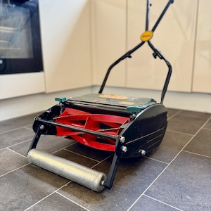 May include: A manual push lawn mower featuring a black frame, a bright red cylindrical cutting blade assembly, and a silver metal front roller. The machine includes a green top cover with the brand name Webb printed in yellow and red text.