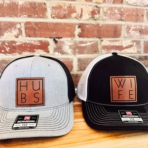 May include: Two black and grey trucker hats with brown leather patches. The patch on the left hat says "HUSB" and the patch on the right hat says "WIFE".