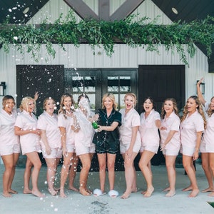 May include: A group of women in matching pink pajamas celebrate with a champagne pop. The women are smiling and laughing as they stand in front of a white building with a green garland.