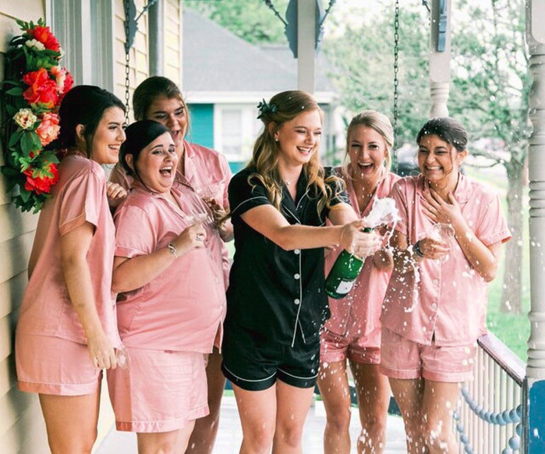 May include: A group of six women in matching pink pajamas celebrate with a bottle of champagne. They are smiling and laughing as they pop the cork. The women are standing on a porch with a floral wreath on the wall behind them.