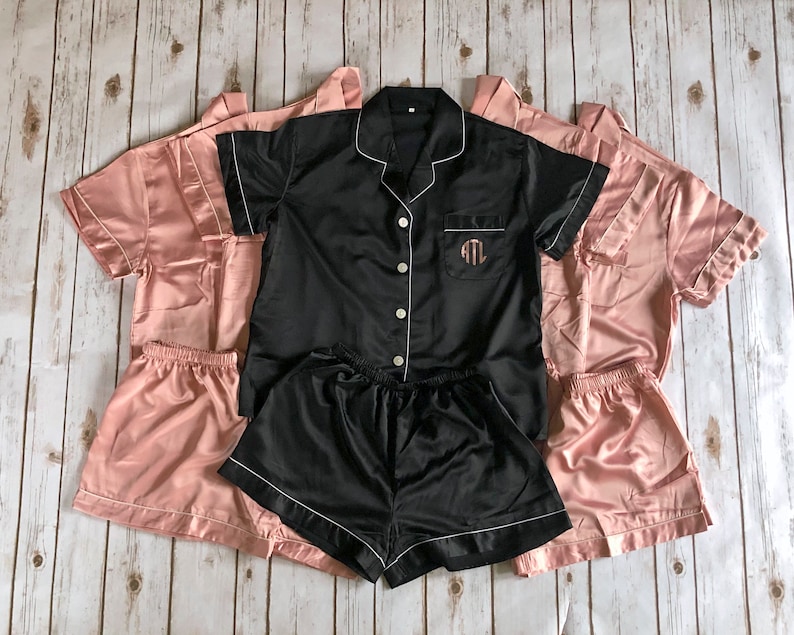 May include: Three pairs of pajamas, one black with white trim and the initials "ATL" embroidered on the chest, and two in a light pink color. All three pairs have short sleeves and shorts.