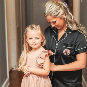 May include: A woman in a black satin robe with white piping and the monogram "MAB" embroidered on the pocket is helping a young girl put on a light brown dress with ruffled sleeves.