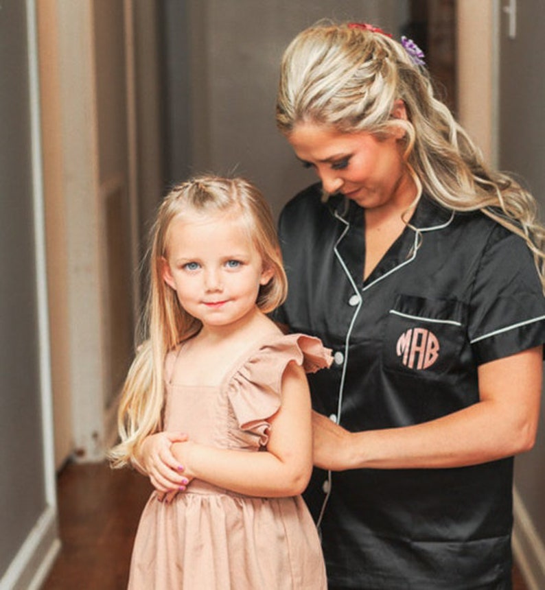 May include: A woman in a black satin robe with white piping and the monogram "MAB" embroidered on the pocket is helping a young girl put on a light brown dress with ruffled sleeves.
