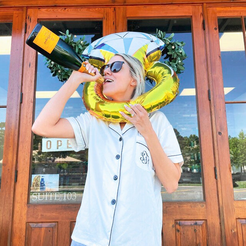 May include: A person wearing a white satin pajama top with black trim and a gold ring on their finger. They are holding a bottle of champagne and wearing a large gold ring balloon around their head. The balloon is shaped like a diamond ring.