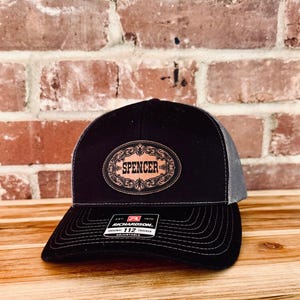 May include: Black trucker hat with a gray mesh back and a curved brim. The hat features a brown oval patch with the name "Spencer" in white script, surrounded by decorative scrollwork. The hat is adjustable.