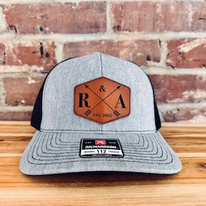 May include: A gray and black trucker hat with a brown leather patch that says "R & A EST. 2025" with crossed arrows.