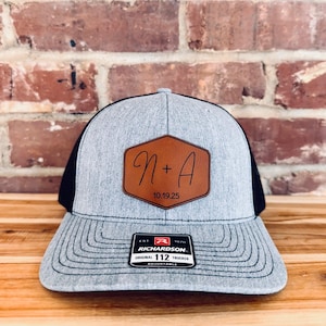 May include: Gray and black trucker hat with a leather patch. The patch features the initials "N + A" and the date "10.19.25". The hat has a curved brim and an adjustable strap. The Richardson brand label is visible.