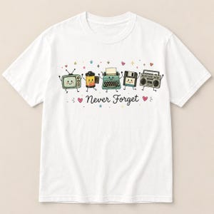 May include: A white t-shirt featuring a graphic of five anthropomorphic vintage items: a television, a film roll, a typewriter, a floppy disk, and a boombox. Below the characters, the text "Never Forget" is printed in black, surrounded by small hearts and stars.