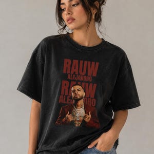 May include: A charcoal black, acid-washed short-sleeve t-shirt featuring a graphic print of Rauw Alejandro. The design includes the text "RAUW ALEJANDRO" in bold red lettering above a photographic image of the artist wearing a red blazer.