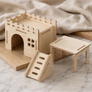 May include: A light-colored wooden small animal habitat set featuring a castle-shaped house with a crenelated roof, an arched doorway, and a four-pane window. The set includes a separate rectangular platform table and a small staircase with four steps.
