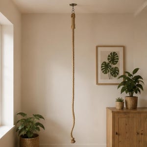 May include: A long, thick, natural-colored twisted jute rope hangs vertically from a dark metal ceiling mount. The rope features a reinforced top section with a metal ring attachment and a knotted end at the bottom, suspended in a room.