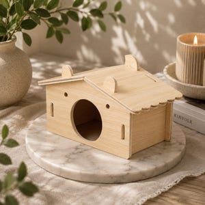 Puede incluir: Una casita de madera de color claro para animales pequeños con un techo a dos aguas que presenta dos protuberancias redondeadas en forma de orejas, una entrada circular y dos pequeños orificios de ventilación redondos sobre la abertura.