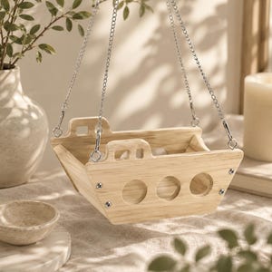 May include: A light-colored wooden hanging planter shaped like a boat, featuring three circular cutouts on the side, a raised cabin section, and silver metal chains attached to the top corners for suspension, showcasing a natural wood grain finish.