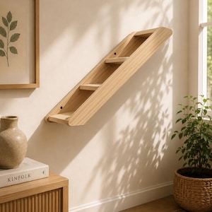 May include: A light-colored wooden wall-mounted cat ladder featuring three horizontal steps set within two parallel side rails. The structure is angled diagonally and attached to the wall, showcasing a natural wood grain finish and visible screw holes.