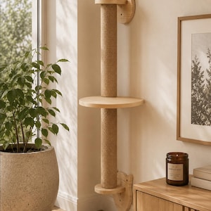 Puede incluir: Una torre de escalada para gatos montada en la pared que cuenta con dos postes rascadores verticales envueltos en sisal, una plataforma circular central de madera y dos soportes de pared de madera con forma de orejas de gato.