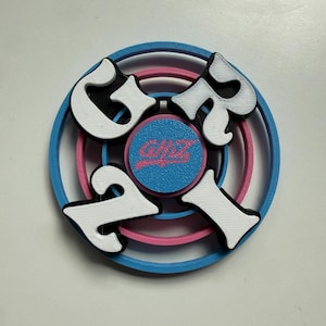 Può includere: Un oggetto circolare stampato in 3D con anelli concentrici blu e rosa. Al centro si trova un piccolo disco rosa con il testo "GHZ" in un carattere stilizzato. Quattro grandi lettere bianche in grassetto formano la parola "GRIP" attorno.