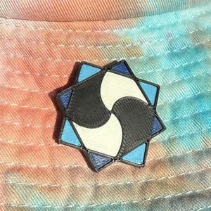 Könnte beinhalten: Eine sternförmige Anstecknadel mit schwarzem Rahmen und einem zentralen Design aus zwei ineinandergreifenden geschwungenen Formen in Schwarz und Weiß, umgeben von acht dreieckigen Abschnitten in abwechselnden Hell- und Dunkelblautönen.