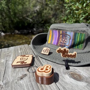 May include: An olive green bucket hat with a rectangular patch featuring vertical stripes and a bread slice logo. Included are a bread-shaped keychain, a small bread slice pin, and a brown bread-shaped clip with the text daily bread.