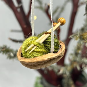 May include: A miniature ornament featuring a walnut shell filled with green yarn and wooden knitting needles. The ornament is suspended by a white ribbon, showcasing a handcrafted, cozy theme.