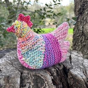 May include: A colorful, hand-knitted chicken figurine. The chicken features a red comb and beak, with a body of blue, purple, pink, and yellow yarn. The knitted hen is sitting on a weathered tree stump, with a blurred green background.