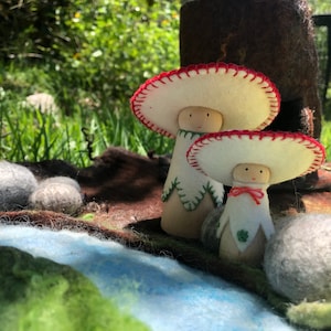 May include: Two felt mushroom dolls with red and white hats and green trim stand on a felt play mat with a blue river and grey rocks.