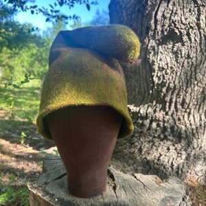 Pode incluir: Um chapéu de feltro verde e castanho com um topo longo e arredondado, exibido numa cabeça de manequim castanha. O chapéu tem uma aparência texturizada e está situado num fundo natural ao ar livre com um tronco de árvore e folhagem.