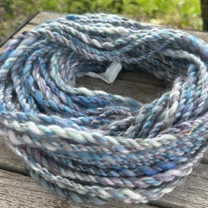 May include: A skein of hand-spun yarn in shades of blue, gray, and lavender. The yarn is thick and textured, with a slight sheen. The fibers are twisted together, creating a unique and artisanal look. Perfect for knitting, crocheting, or other fiber arts projects.
