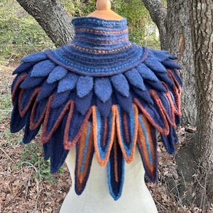 May include: A handcrafted, blue and brown knitted shawl with a feathered design. The shawl features a high collar and layered, petal-like elements in shades of navy, brown, and orange. The piece is displayed on a mannequin, showcasing its intricate texture and design.
