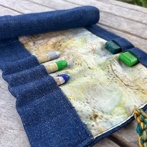 May include: A blue denim roll-up case with a fabric lining and multiple pockets for holding art supplies. The case is rolled up and tied with a green and yellow cord.
