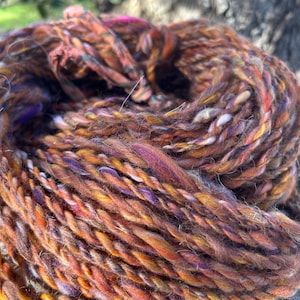 May include: A close-up of a skein of hand-spun yarn. The yarn is a mix of brown, orange, purple, and white fibers. The yarn has a textured appearance, with visible twists and variations in color. The yarn is likely used for knitting or other fiber arts.