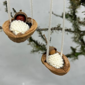 Könnte beinhalten: Zwei handgefertigte Ornamente mit einer Walnussschale als Wiege mit einem weißen, gestrickten Baby darin. Jede Wiege ist mit einem weißen Band aufgehängt, mit einer Eichelkappe als Kopfstütze. Der Hintergrund ist eine verschwommene Ansicht eines Astes.