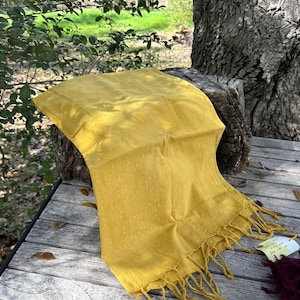 May include: A golden yellow scarf with fringe detail. The scarf is draped over a wooden surface, with a tag attached. The scarf has a dotted pattern and is made of a soft, flowing material. The scarf is a fashion accessory.