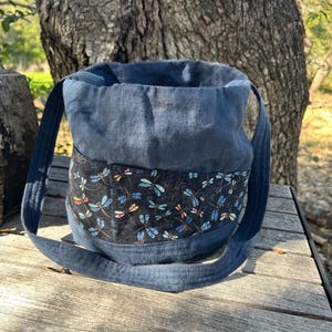 May include: A blue fabric tote bag with a black band featuring a dragonfly pattern. The bag has a drawstring closure and long straps for carrying over the shoulder. The bag is made of a sturdy fabric and is suitable for everyday use.
