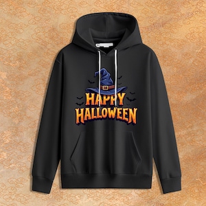 Puede incluir: Una sudadera negra con capucha que presenta un gráfico de un sombrero de bruja azul con un cinturón marrón, rodeado de pequeños murciélagos negros. Debajo del sombrero, el texto "HAPPY HALLOWEEN" está impreso en letras naranjas degradadas.