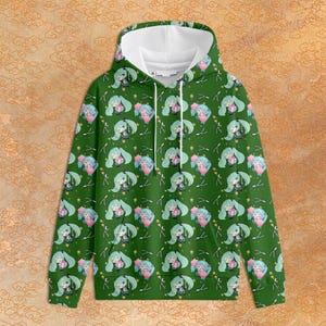 Puede incluir: Una sudadera con capucha verde de manga larga con un patrón repetitivo de un personaje de anime de pelo verde azulado sosteniendo un regalo rosa, intercalado con pequeñas estrellas blancas y líneas finas, con forro interior blanco.